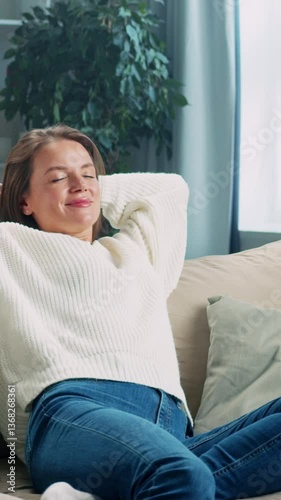 Young woman is sitting on a couch in a living room. She is relaxing and enjoying her free time. The video is in slow motion