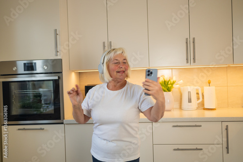 Obraz na płótnie A joyful senior woman stands in her kitchen, taking a selfie with her phone. She smiles happily, capturing a moment of enjoyment. A beautiful setting with modern kitchen appliances w salonie