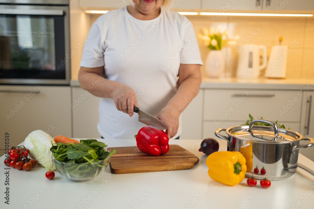 A woman is chopping a fresh red bell pepper on a wooden cutting board in the kitchen. Surrounding her are carrots, green leaves, and a saucepan with fresh vegetables