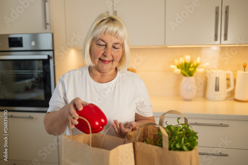 Obraz na płótnie An elderly woman is unpacking groceries from paper bags in her kitchen. She places a jar of food and fresh produce on the counter, enjoying the convenience of home delivery w salonie