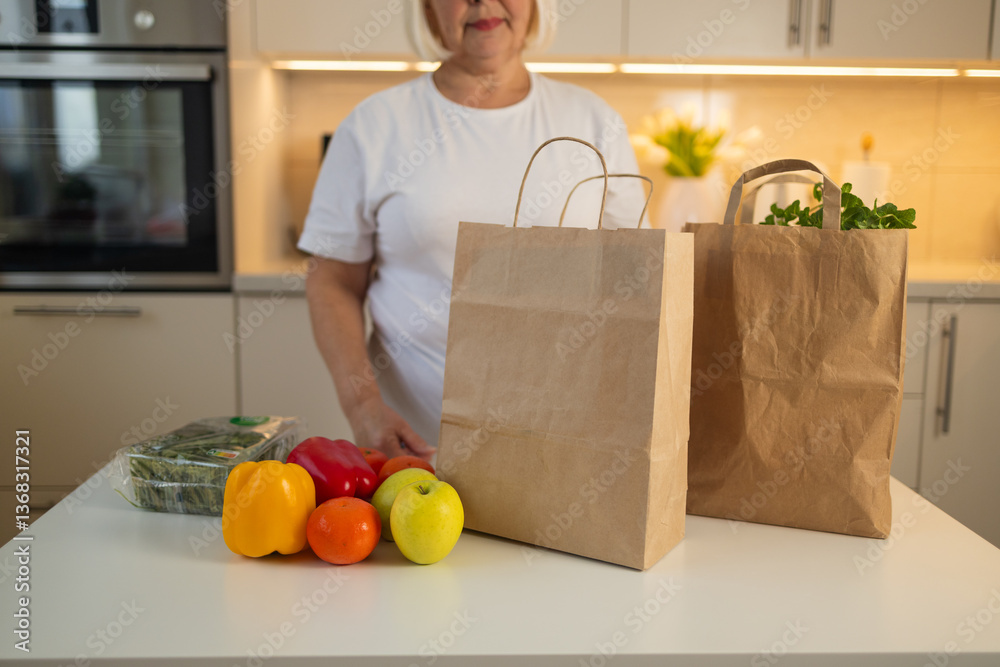 An elderly woman is standing in her kitchen with fresh groceries, including colorful bell peppers and apples, on the table. She is surrounded by brown paper grocery bags.