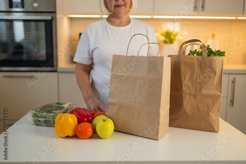 Obraz na płótnie An elderly woman is standing in her kitchen with fresh groceries, including colorful bell peppers and apples, on the table. She is surrounded by brown paper grocery bags. w salonie