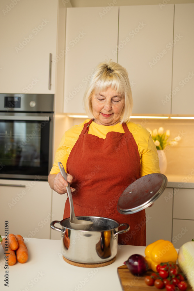 A senior woman in a yellow apron is stirring vegetables in a pot on the kitchen counter, with fresh carrots, tomatoes, and other ingredients around her.