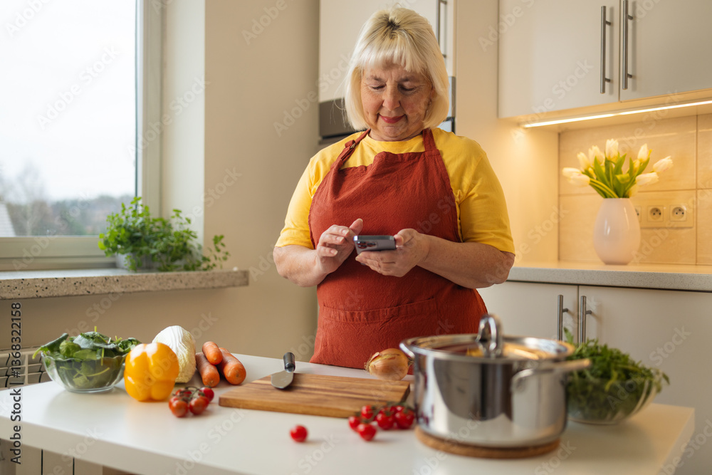 A woman in a yellow shirt and apron is preparing food in the kitchen. She looks at her phone for a recipe while chopping vegetables like tomatoes and bell peppers.