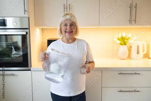 A happy senior woman holding a water pitcher and a glass in the kitchen, ready to pour herself a refreshing drink. This image reflects healthy habits and enjoying moments at home