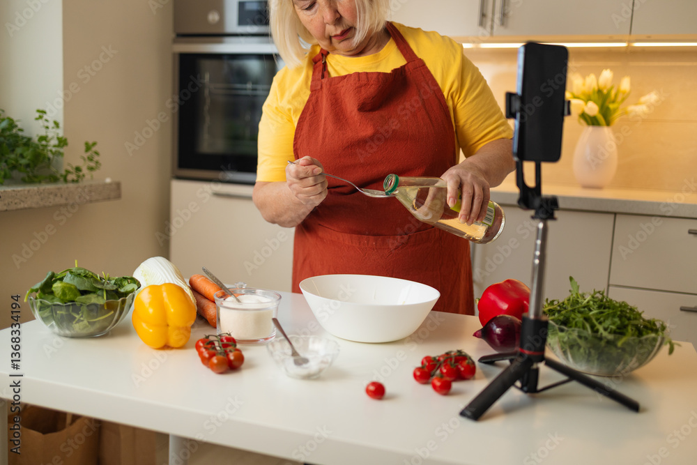 A senior woman in a yellow shirt and brown apron adds vinegar to a bowl as she prepares a meal in the kitchen. Fresh vegetables and kitchen tools are neatly arranged on the counter.