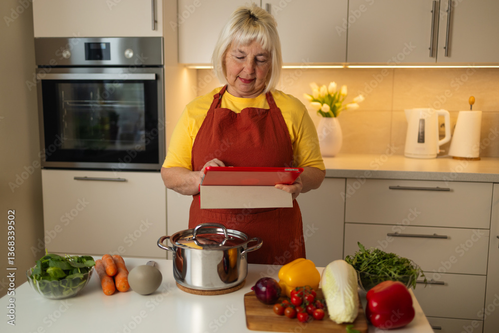 A senior woman in a yellow shirt and red apron stands in her kitchen, looking at a tablet while preparing vegetables like carrots, tomatoes, and cabbage for cooking.
