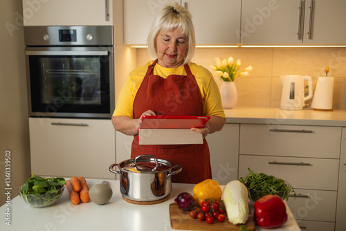 Obraz na płótnie A senior woman in a yellow shirt and red apron stands in her kitchen, looking at a tablet while preparing vegetables like carrots, tomatoes, and cabbage for cooking. w salonie