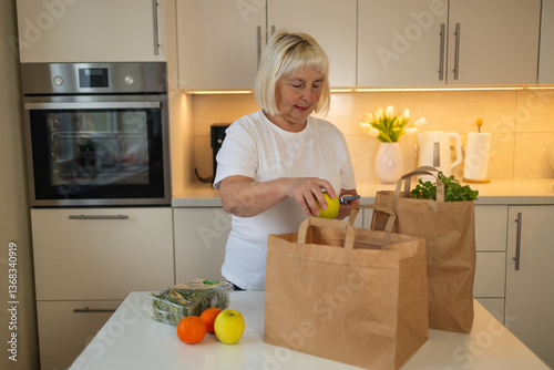 An elderly woman is unpacking groceries from paper bags in her kitchen. She places a jar of food and fresh produce on the counter, enjoying the convenience of home delivery