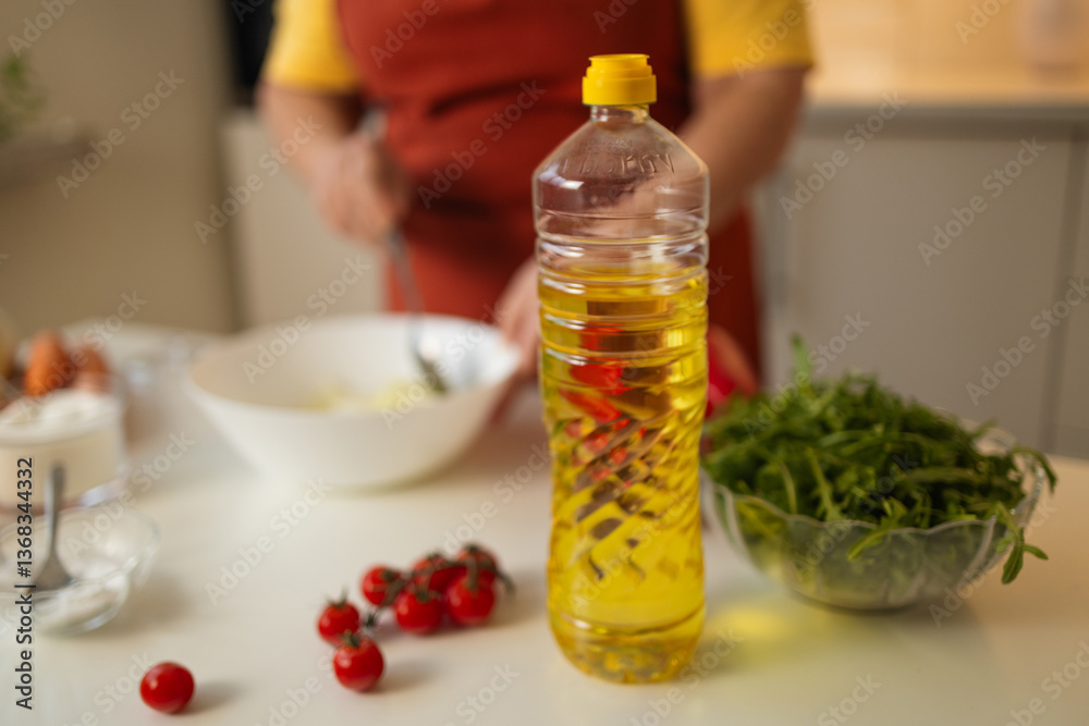 A bottle of cooking oil stands on the kitchen counter, surrounded by fresh vegetables like tomatoes and greens, while a senior woman prepares a dish in the background