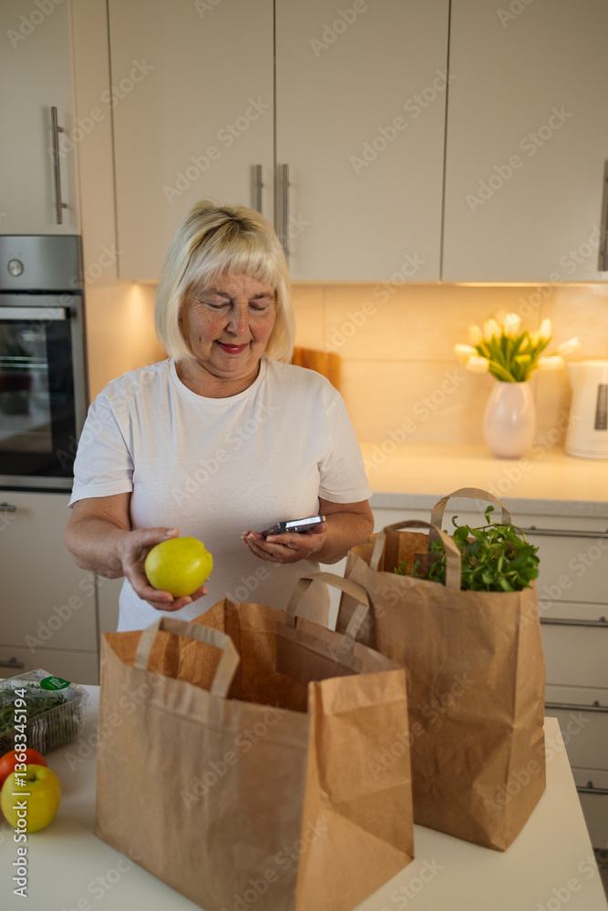 An elderly woman is unpacking groceries from paper bags in her kitchen. She places a jar of food and fresh produce on the counter, enjoying the convenience of home delivery