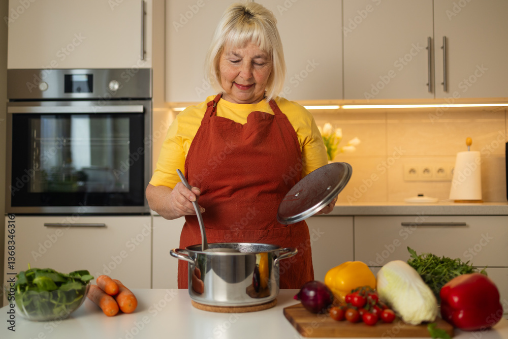 A senior woman in a yellow apron is stirring vegetables in a pot on the kitchen counter, with fresh carrots, tomatoes, and other ingredients around her.