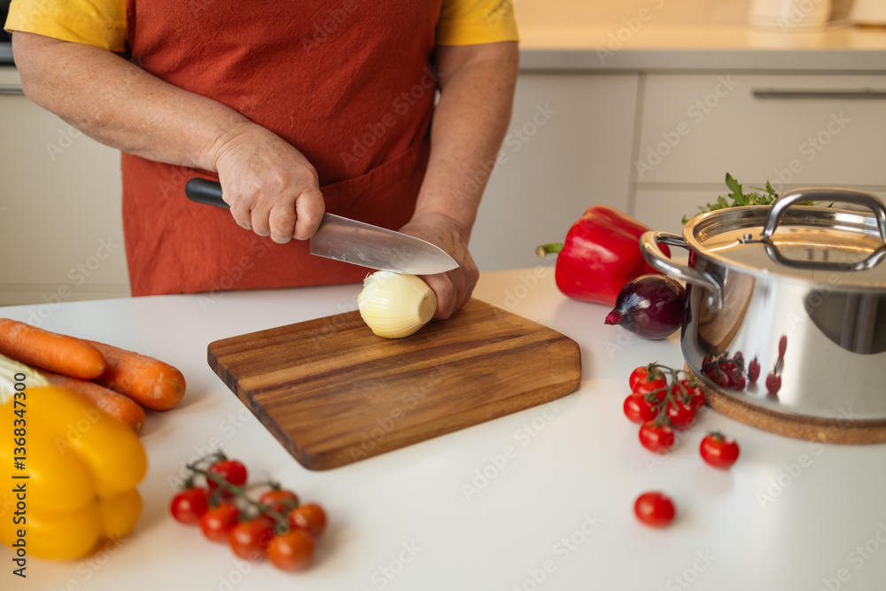 A woman in a yellow shirt and apron is peeling an onion in the kitchen. She is preparing ingredients for a meal while surrounded by fresh vegetables like carrots, tomatoes, and bell peppers