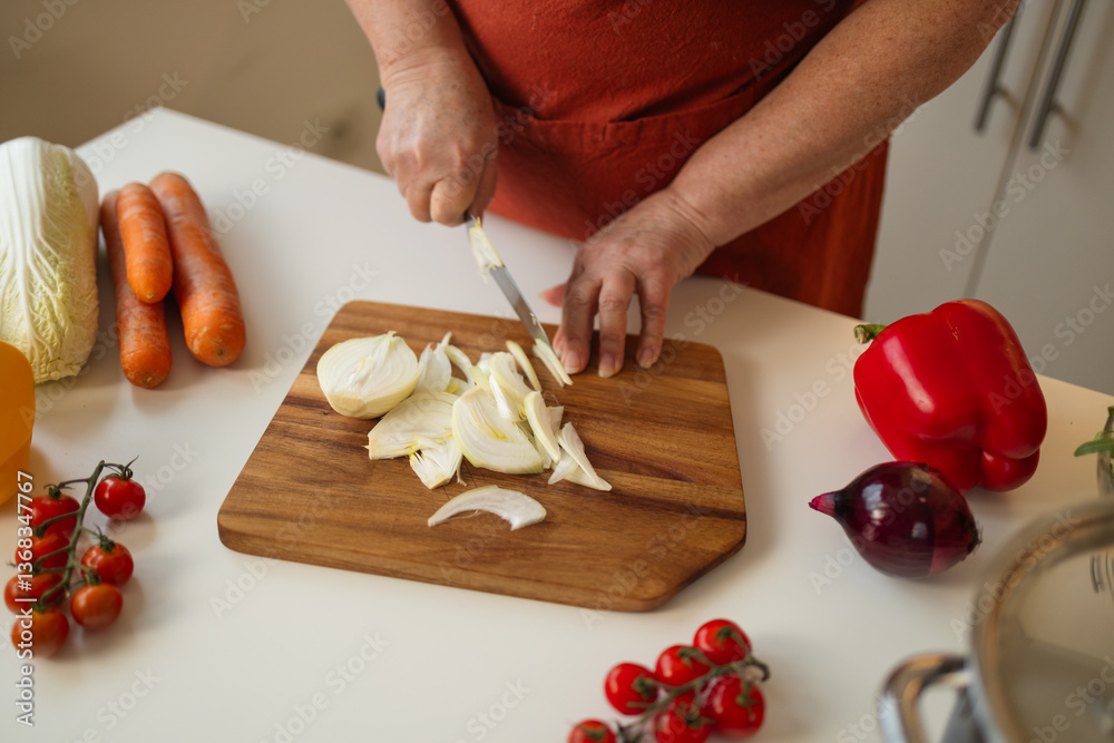 A person chopping onions on a wooden cutting board in a kitchen. Nearby, various fresh vegetables such as tomatoes, carrots, and peppers are spread out, preparing for cooking