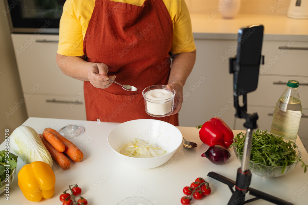 A senior woman in a yellow shirt and brown apron adds sugar to a bowl while preparing a meal in a well-organized kitchen. Fresh vegetables and kitchen tools are visible on the countertop