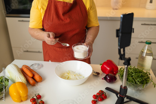Obraz na płótnie A senior woman in a yellow shirt and brown apron adds sugar to a bowl while preparing a meal in a well-organized kitchen. Fresh vegetables and kitchen tools are visible on the countertop w salonie