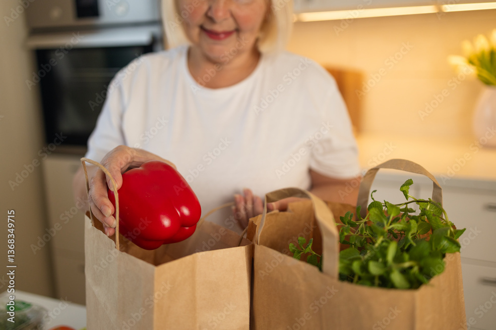 An elderly woman is unpacking groceries from paper bags in her kitchen. She places a jar of food and fresh produce on the counter, enjoying the convenience of home delivery