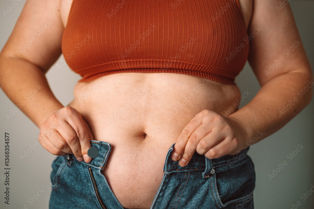 A woman trying to button up tight jeans, symbolizing challenges with body image and weight issues. This reflects the daily struggles of self-image and fitting into clothes, common among many
