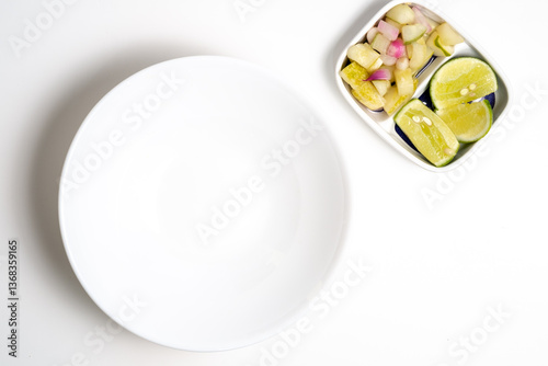A white, empty round plate sits beside a small, angled dish containing Indonesian pickles (acar) of sliced cucumbers, onions, and green chili, plus lime wedges. Minimalist composition. photo