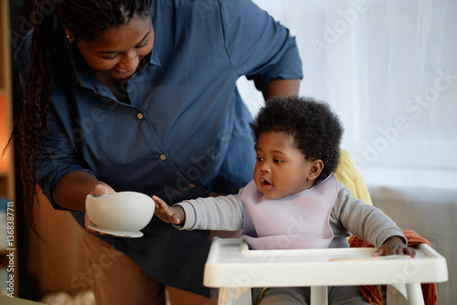 Medium shot of hungry Black baby girl reaching out to take plastic bowl with tasty fruit puree from mothers hands, while sitting in high chair during mealtime in kitchen