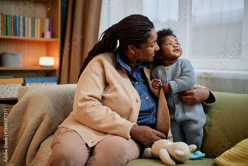 Medium full shot of happy Black woman wearing comfy cardigan gently hugging cute little baby girl dressed in knitted romper of blue color captured together on couch at home, heartfelt bonding moment
