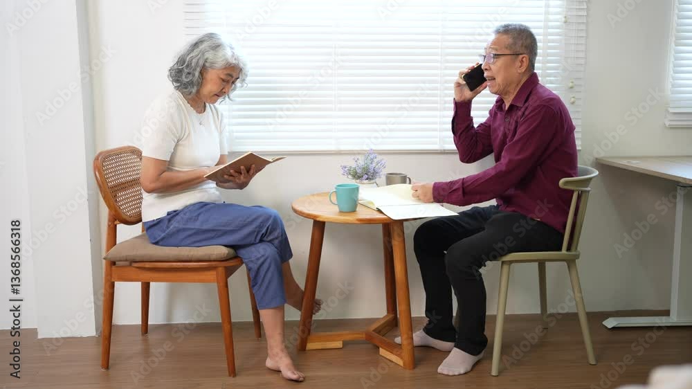 Happy elderly couple sitting at wooden table, cheerful man talking on phone while senior woman reads book in cozy home setting. Happy elderly couple sitting at wooden table, cheerful man talking on phone while senior woman reads book in cozy home setting.