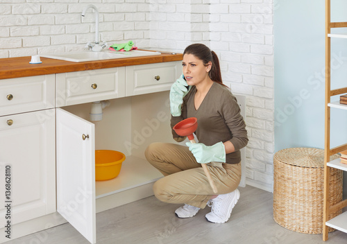 Young girl talking by the phone advising with plumber how to fix broken pipe in kitchen or unclog sink drain. Woman trying to clean sink using plunger at home. Housekeeping and household work concept