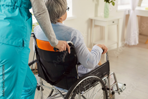 Nurse or health care worker in uniform pushing disabled female patient in wheelchair in medical clinic. Handicapped woman with disability in retirement home. Rehab and caregiver support concept.