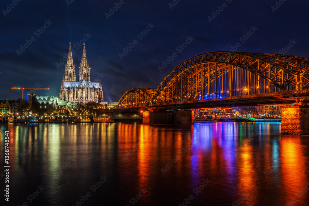 Fototapeta premium As dusk descends over Cologne, the blue hour transforms the city into an enchanting spectacle. In this photograph, the Hohenzollernbrücke, with its robust arches and love locks, forms a powerful foreg