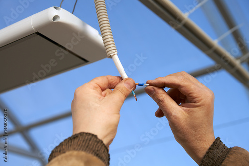 Installation of infrared heater to the ceiling of polycarbonate greenhouse with electric cable in corrugated. Male hands with wire terminals.