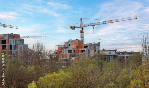Obraz na płótnie A modern residential complex under construction, featuring concrete and brick structures with large glass windows, surrounded by trees, with cranes towering above against a clear blue sky. w salonie