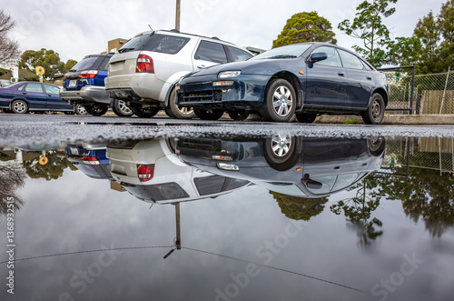 A row of parked cars is reflected in a large puddle on wet asphalt after rainfall or storm, indicating water accumulation and drainage issues in an urban city or parking lot.