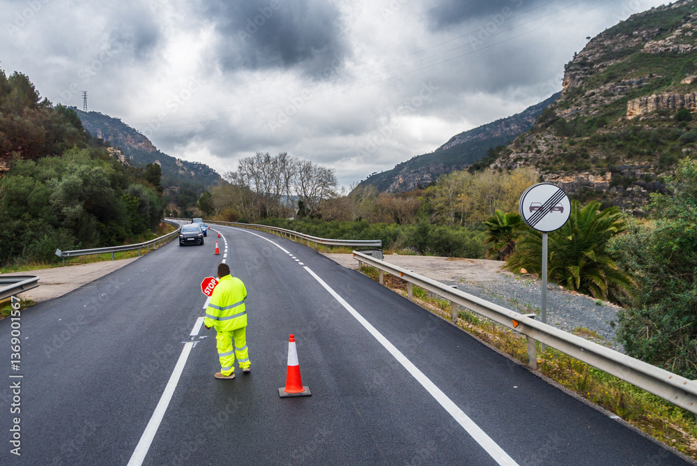 Road maintenance worker in the center of the roadway, closing one lane to allow alternating traffic on a two-way road under construction.
