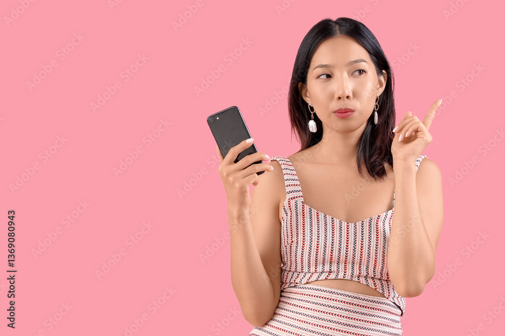 Thoughtful young beautiful Asian woman with mobile phone pointing at something on pink background Thoughtful young beautiful Asian woman with mobile phone pointing at something on pink background