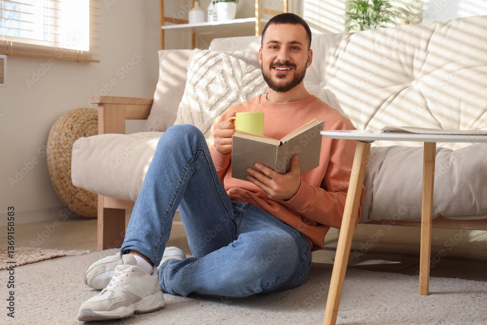Handsome young man with cup of tea reading book while sitting on floor in living room Handsome young man with cup of tea reading book while sitting on floor in living room