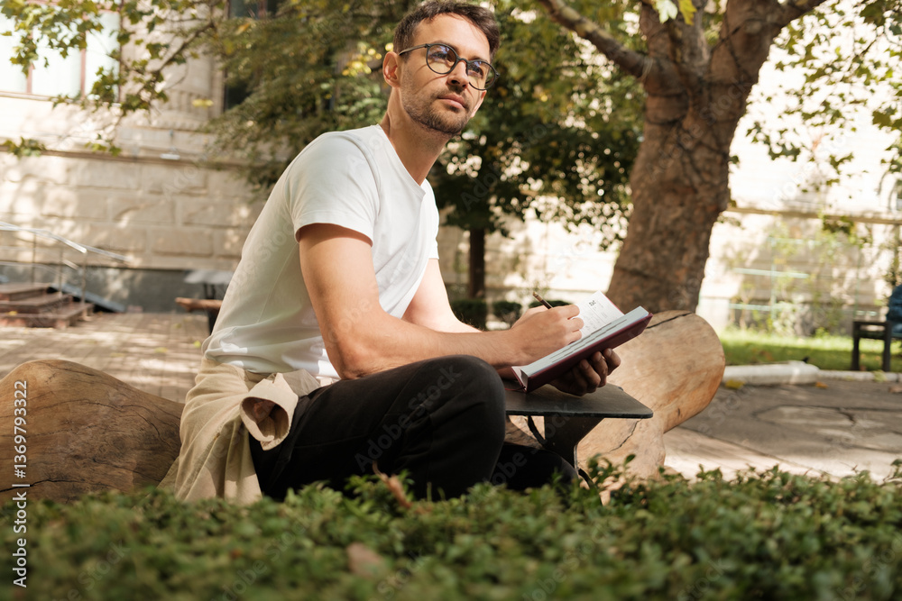 A thoughtful man with glasses sits on a log in a park, diligently writing in his notebook, surrounded by greenery and sunlight. The scene conveys creativity, reflection, and relaxation.