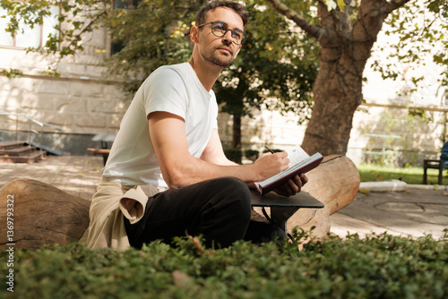 Obraz na płótnie A thoughtful man with glasses sits on a log in a park, diligently writing in his notebook, surrounded by greenery and sunlight. The scene conveys creativity, reflection, and relaxation. w salonie
