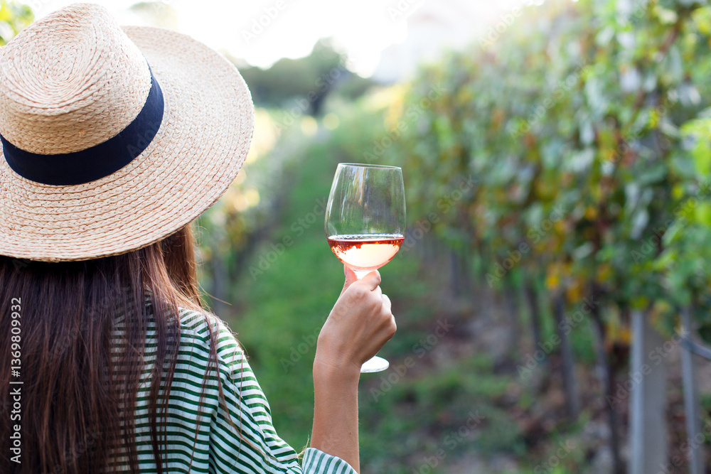 Obraz premium Woman with wineglass of pink wine in vineyard outdoors. Traveler tasting alcohol-free wine in winery. Female farmer in straw hat relaxing, enjoying harvest. Authentic small business, local shop