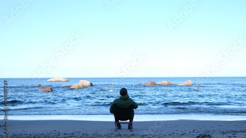 A man sits on a chair facing the sea and drinks from a mug