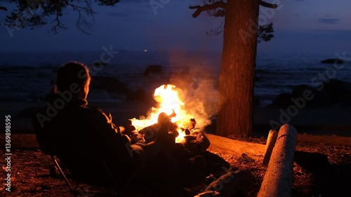 A man sits at night in front of a fire and drinks. Against the backdrop of the sea