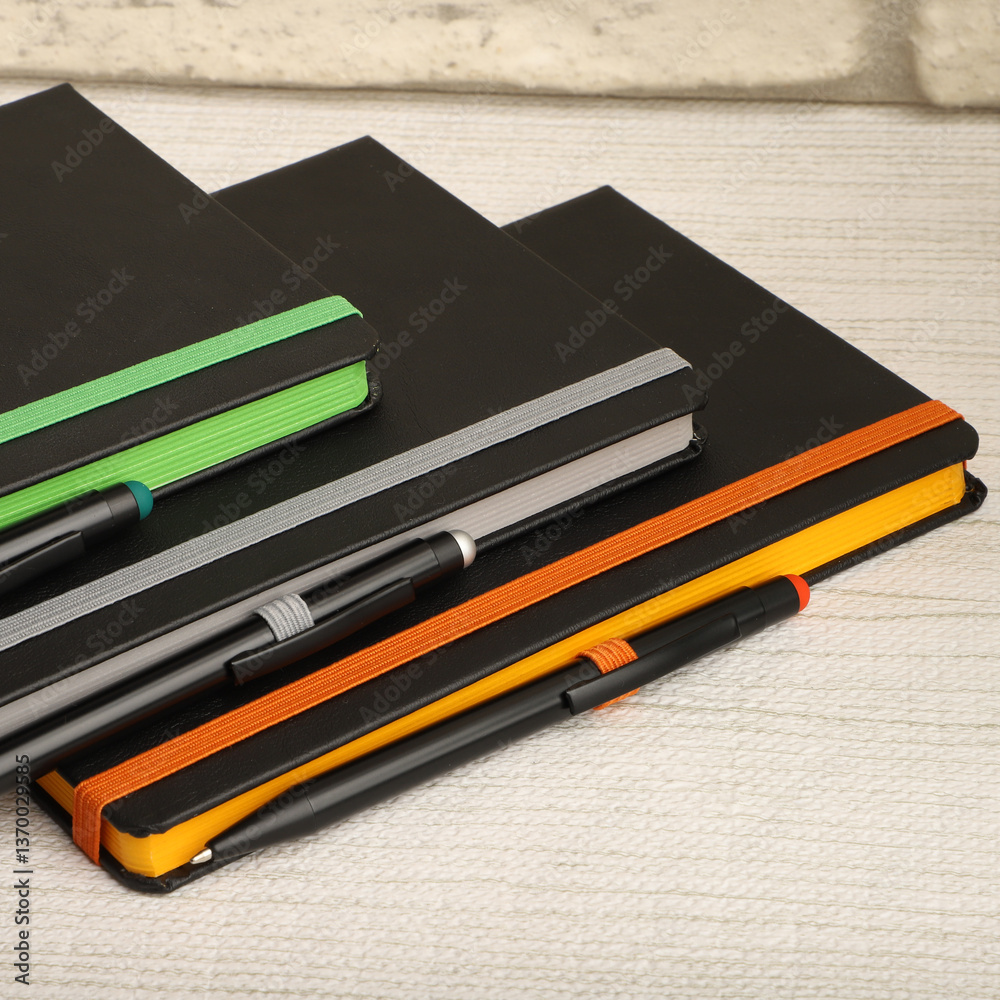 Black notebooks with colorful elastic bands and pens resting on textured surface