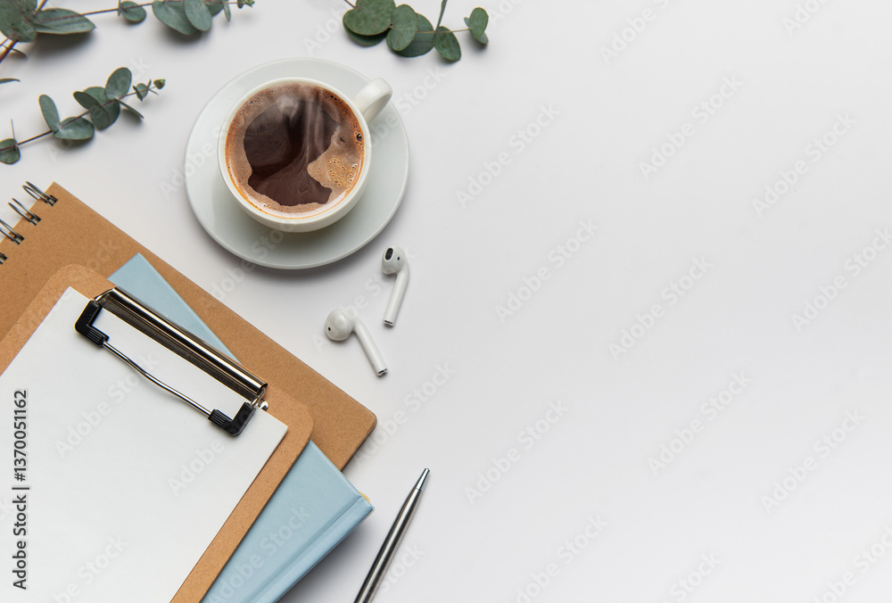 Eucalyptus, coffee cup, clipboard, notebook, pen, and wireless headphones resting on white desk