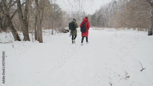 Two people walking through a snowy forest in winter, surrounded by tall trees and a serene atmosphere