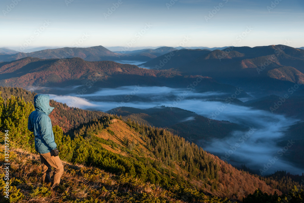 Fototapeta premium Rear view of a young man wearing a blue jacket, gazing at the breathtaking view from the top of a mountain at sunrise, with low clouds filling the valley below