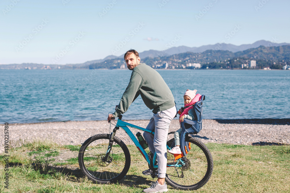 Fototapeta premium A father riding a bicycle with his young child seated in a safety seat on the back. They are enjoying a scenic outdoor ride near a beautiful lake with mountains in the background on a sunny day