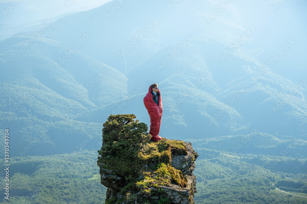 Obraz premium Young European man standing in a red sleeping bag on the edge of a rocky mountain peak, surrounded by mountain ridges.