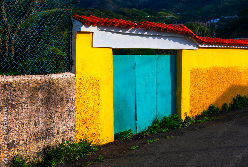 Bright yellow wall paired with a turquoise gate, crowned by a tiled roof. Vibrant teal doors stand out against a backdrop of sunny yellow walls, creating a striking visual contrast