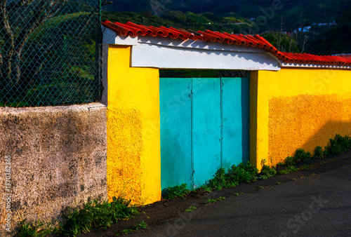 Obraz na płótnie Bright yellow wall paired with a turquoise gate, crowned by a tiled roof. Vibrant teal doors stand out against a backdrop of sunny yellow walls, creating a striking visual contrast w salonie