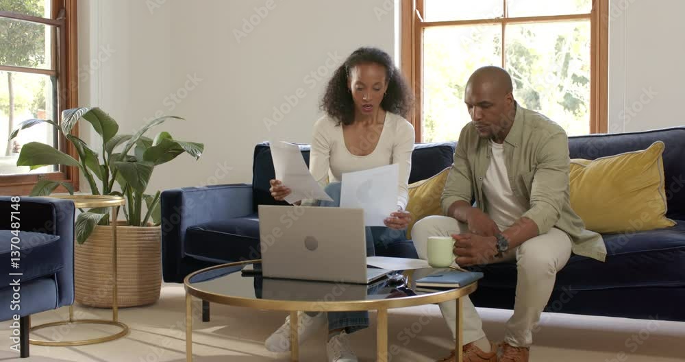 Happy diverse couple sitting on sofa, using laptop and enjoying conversation at home