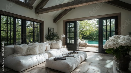 Cozy living room with white plush seating dark framed windows wooden beams and doors opening to greenery and a pool with white flower arrangements